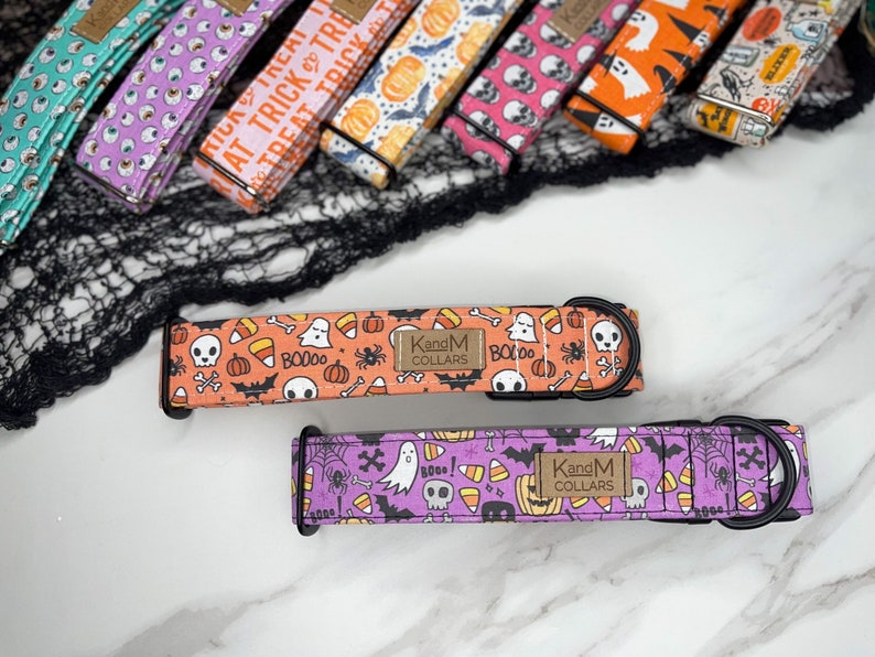May include: Two Halloween-themed dog collars with orange and purple backgrounds. The collars feature a variety of spooky designs, including ghosts, skulls, bats, candy corn, and pumpkins. The collars are made by KandM Collars.
