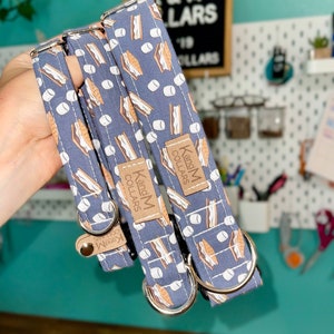 May include: Three dog collars with a blue background and a pattern of brown graham crackers, white marshmallows, and chocolate bars. The collars have silver metal hardware and a brown leather tag that reads "K and M Collars".
