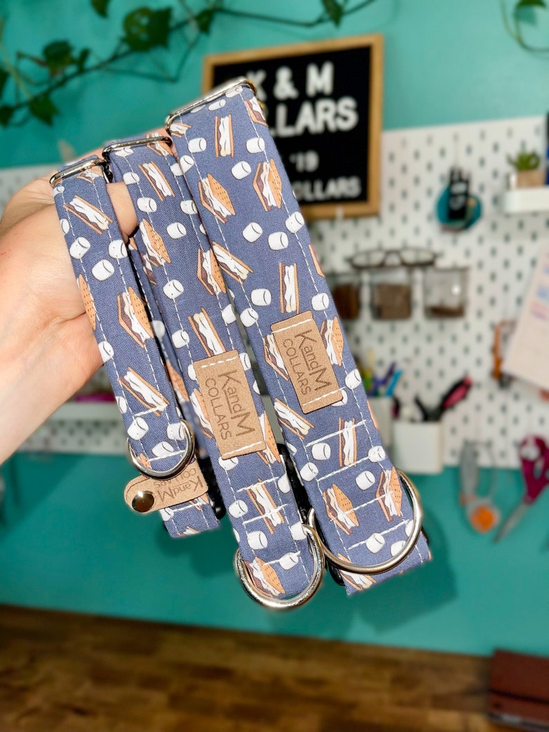 May include: Three dog collars with a blue background and a pattern of brown graham crackers, white marshmallows, and chocolate bars. The collars have silver metal hardware and a brown leather tag that reads "K and M Collars".