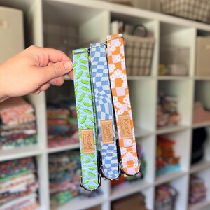 May include: Three fabric pet collars featuring distinct patterns: a light blue collar with green pickle shapes, a blue and white wavy checkerboard collar, and an orange and white checkerboard collar with white flowers. Each has a tan leather tag.