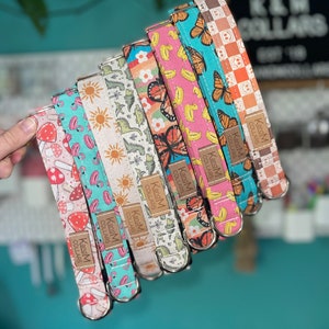 May include: A stack of eight colorful dog collars with different patterns, including mushrooms, flamingos, butterflies, dinosaurs, and bananas. Each collar has a silver buckle and a brown leather tag that reads "KandM Collars".