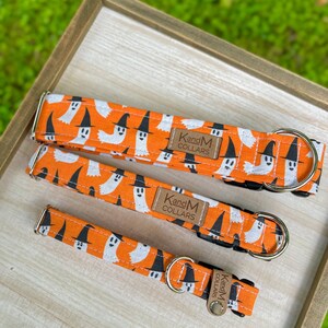 May include: Three orange and white dog collars with a ghost and witch hat pattern. The collars have black webbing and brown leather labels that say "KandM Collars".