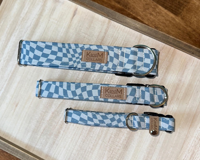 May include: Three pet collars in varying sizes featuring a light blue and white wavy checkerboard pattern. Each collar includes a black plastic buckle, a silver metal D-ring, and a tan leather-like rectangular tag embossed with KandM COLLARS.
