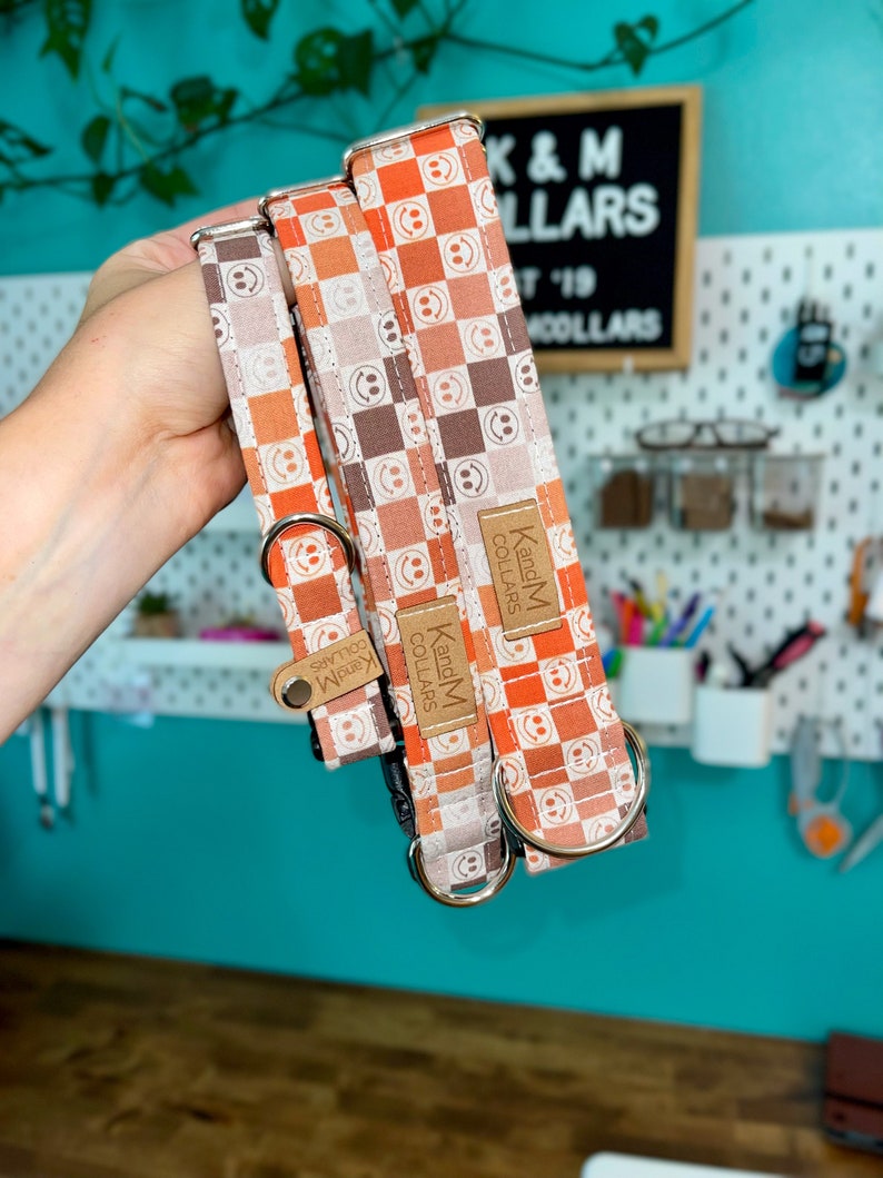 May include: Three dog collars with a checkered pattern in shades of orange, brown, and white. The collars feature a smiley face design and have silver hardware. The collars are labeled "K and M Collars".