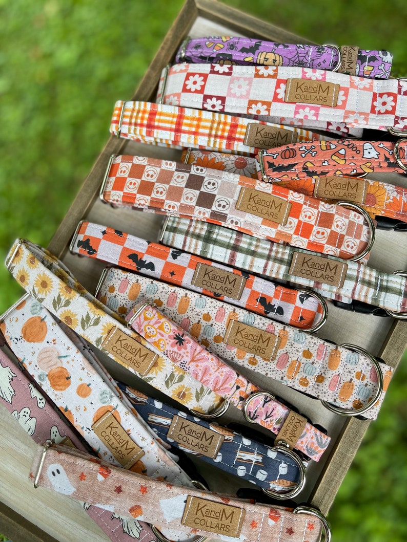 May include: A stack of colorful dog collars with various fall-themed patterns, including pumpkins, ghosts, and plaid. Each collar has a brown leather tag with the text "KandM Collars".