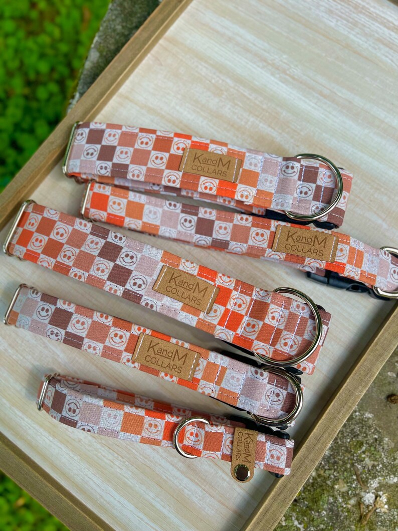 May include: Five dog collars in different sizes with a checkered pattern featuring orange, brown, and white smiley faces. The collars are made by "KandM Collars".