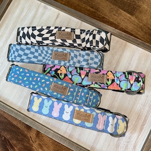 May include: Five fabric pet collars with metal D-rings and buckles. Patterns include black and white checkerboard, blue checkerboard, colorful mushrooms on black, blue with multi-colored jellybeans, and blue with pastel bunnies. Each has a KandM COLLARS tag.