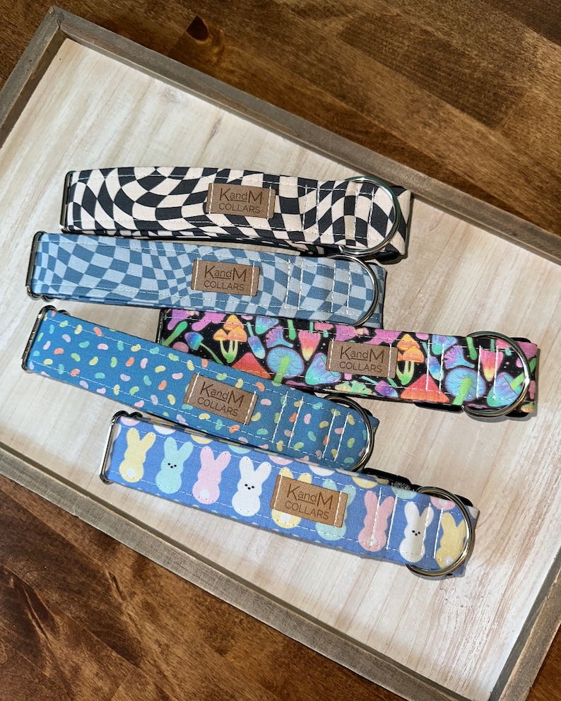 May include: Five fabric pet collars with metal D-rings and buckles. Patterns include black and white checkerboard, blue checkerboard, colorful mushrooms on black, blue with multi-colored jellybeans, and blue with pastel bunnies. Each has a KandM COLLARS tag.