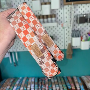 May include: Two orange and white dog collars with a checkered pattern and smiley faces. The collars have silver hardware and a brown leather tag that reads "KandM Collars".