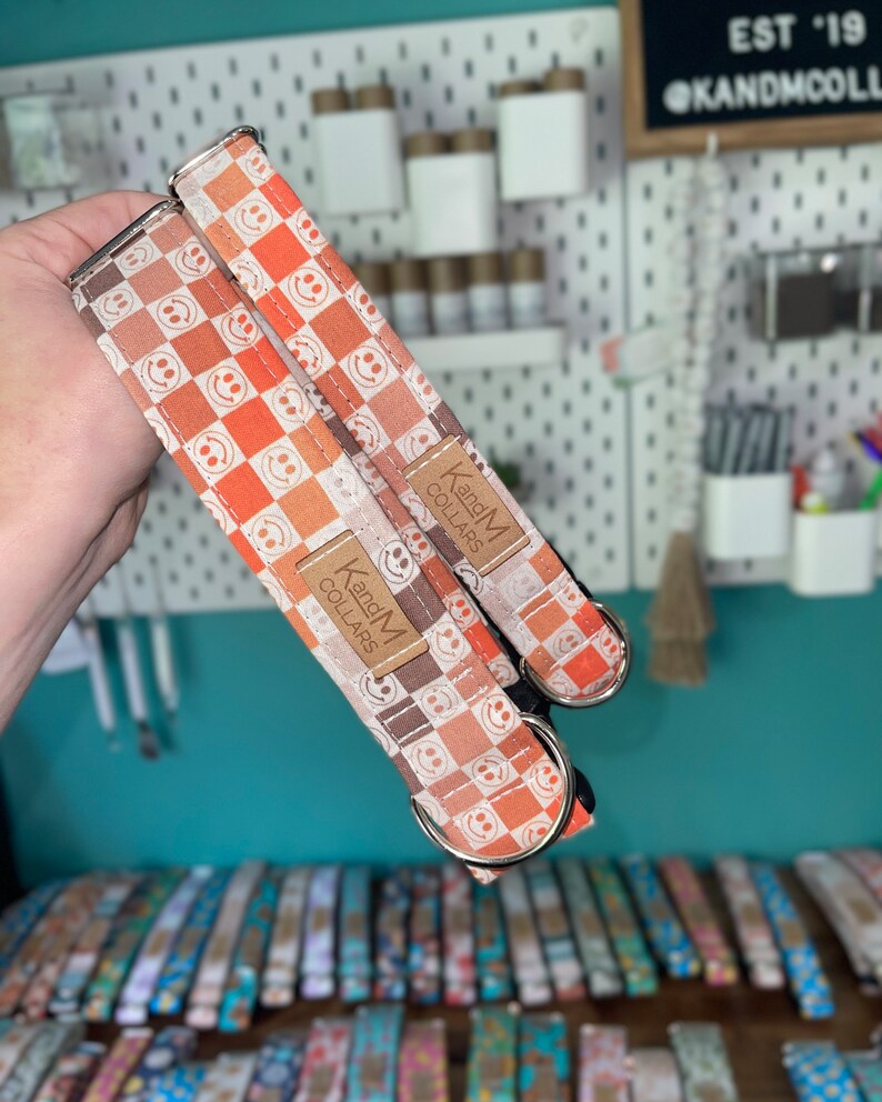 May include: Two orange and white dog collars with a checkered pattern and smiley faces. The collars have silver hardware and a brown leather tag that reads "KandM Collars".