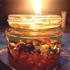 Könnte beinhalten: Eine Kerze im klaren Glasgefäß mit brennender Flamme, die eine Mischung aus rotem und orangefarbenem strukturiertem Wachs, dunklen botanischen Stücken und kleinen hellen Einschlüssen enthält, die in das durchscheinende Wachs eingebettet sind.