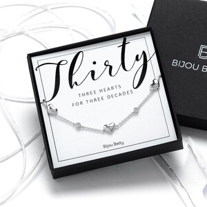 May include: A silver necklace with three heart charms and the text "Thirty Three Hearts for Three Decades" on a white card in a black box. The box has the text "Bijou Betty" on it.