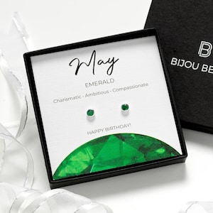 May include: Emerald stud earrings in a black gift box, with the text "May Emerald" and "Happy Birthday!" The open box displays the earrings. The closed box has the brand name "Bijou Betty". Ribbons decorate the scene.