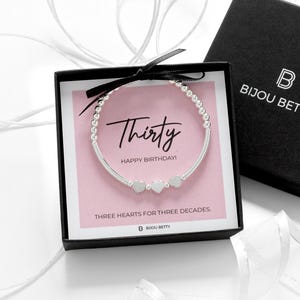 Könnte beinhalten: Ein silbernes Armband mit Herzanhängern wird in einer schwarzen Geschenkbox mit einer rosa Karte mit der Aufschrift "Thirty Happy Birthday!" und "Three Hearts for Three Decades" präsentiert. Die Box ist von Bijou Betty.