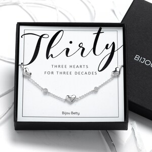 May include: A silver necklace with three heart charms and the text "Thirty Three Hearts for Three Decades" in a black box with the text "Bijou Betty".