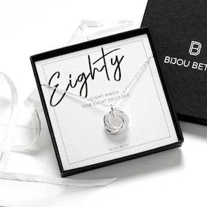 May include: A silver necklace with eight interlocking rings presented in a black gift box. The box insert features the text Eighty, EIGHT RINGS FOR EIGHT DECADES, and BIJOU BETTY. The lid of the box is black with the white logo BIJOU BETTY.
