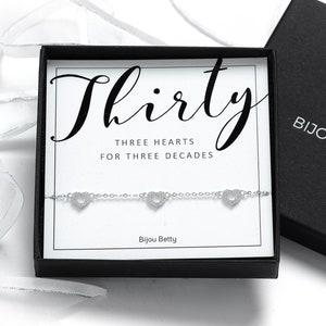 May include: A silver bracelet with three heart charms in a black box. The box has a white label with the text "Thirty Three Hearts For Three Decades" and the logo "Bijou Betty".