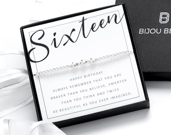 May include: A silver bracelet with small decorative elements is displayed on a card that reads "Sixteen" and "Happy Birthday." The card is inside a black box with the "Bijou Betty" logo. White ribbon and a second black box are also visible.