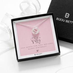 May include: A silver necklace with five interlocking rings, displayed in a black box with the brand name "BIJOU BETTY". The box is open, showing a pink card with the word "Fifty" and the message "FIVE RINGS FOR FIVE DECADES HAPPY BIRTHDAY!"