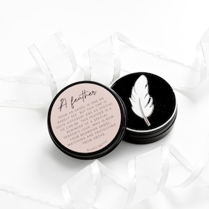 May include: Two small, round, black tins with white feather designs. One tin has a light pink label with the text "A feather" and a quote. White ribbon decorates the background.