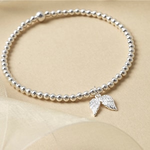 May include: A silver beaded bracelet featuring a charm of two angel wings. The bracelet is composed of small, round, silver beads. The wings charm has etched detailing. The bracelet is presented on a cream-colored, silky surface.
