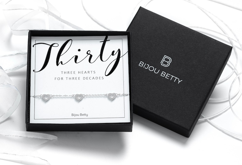 May include: A silver bracelet with three heart charms in a black box. The box has a white label with the text "Thirty Three Hearts For Three Decades" and the logo "Bijou Betty".