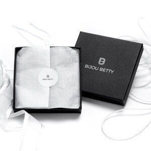 May include: A black gift box with a white logo that says "Bijou Betty" and a white "B" inside a square. The box is open and filled with white tissue paper. A white ribbon is tied around the box.