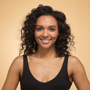 May include: A woman with curly dark brown hair is smiling at the camera. She is wearing a black tank top and a silver necklace with a small pendant.