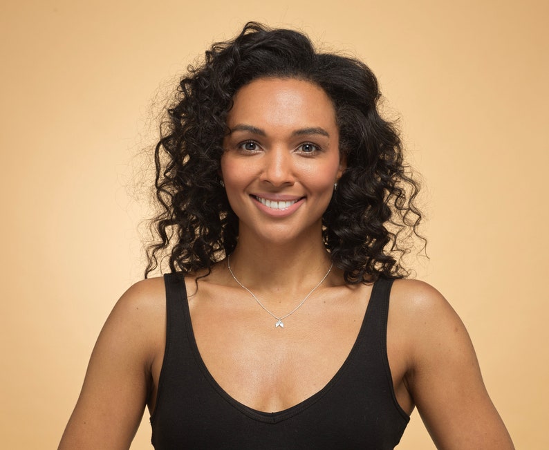 May include: A woman with curly dark brown hair is smiling at the camera. She is wearing a black tank top and a silver necklace with a small pendant.