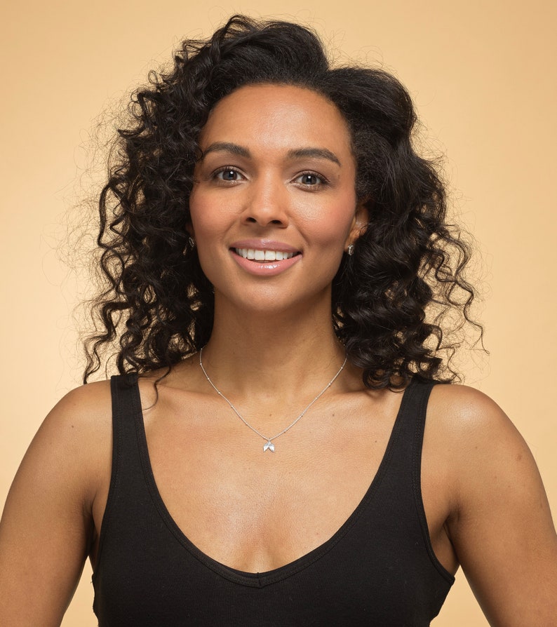 May include: A woman with curly dark brown hair wearing a black tank top and a silver necklace with a small pendant. She is smiling and looking at the camera.