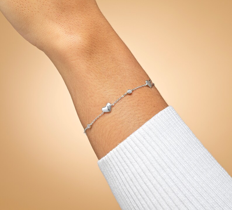 May include: A silver bracelet with a delicate chain and small heart charms. The bracelet has a single clear gemstone accent.