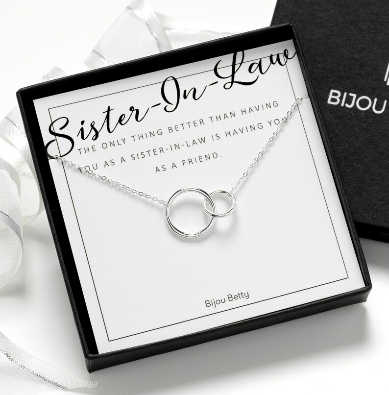 May include: A silver necklace with two interlocking circular rings displayed on a white card inside a black gift box. The card features the text: Sister-In-Law, The only thing better than having you as a sister-in-law is having you as a friend.