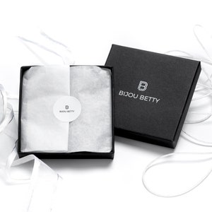 May include: A black gift box with a white logo that says "Bijou Betty" and a white "B" inside a square. The box is open and filled with white tissue paper. A white ribbon is tied around the box.