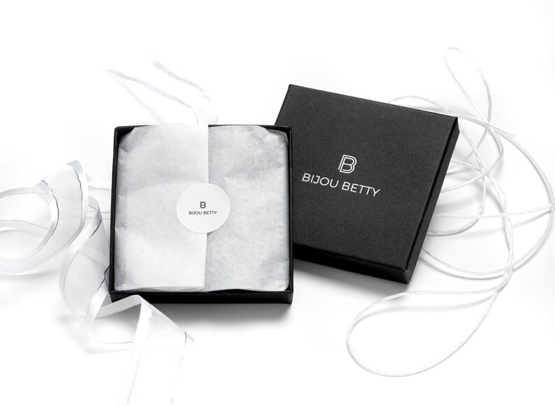 May include: A black gift box with a white logo that says "Bijou Betty" and a white "B" inside a square. The box is open and filled with white tissue paper. A white ribbon is tied around the box.