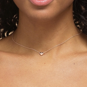 K&ouml;nnte beinhalten: Eine zarte goldene Halskette mit einem kleinen Herzanh&auml;nger. Der herzf&ouml;rmige Anh&auml;nger ist auf einer d&uuml;nnen Kette zentriert, wodurch ein minimalistisches und elegantes Design entsteht. Die Halskette wird um den Hals getragen.