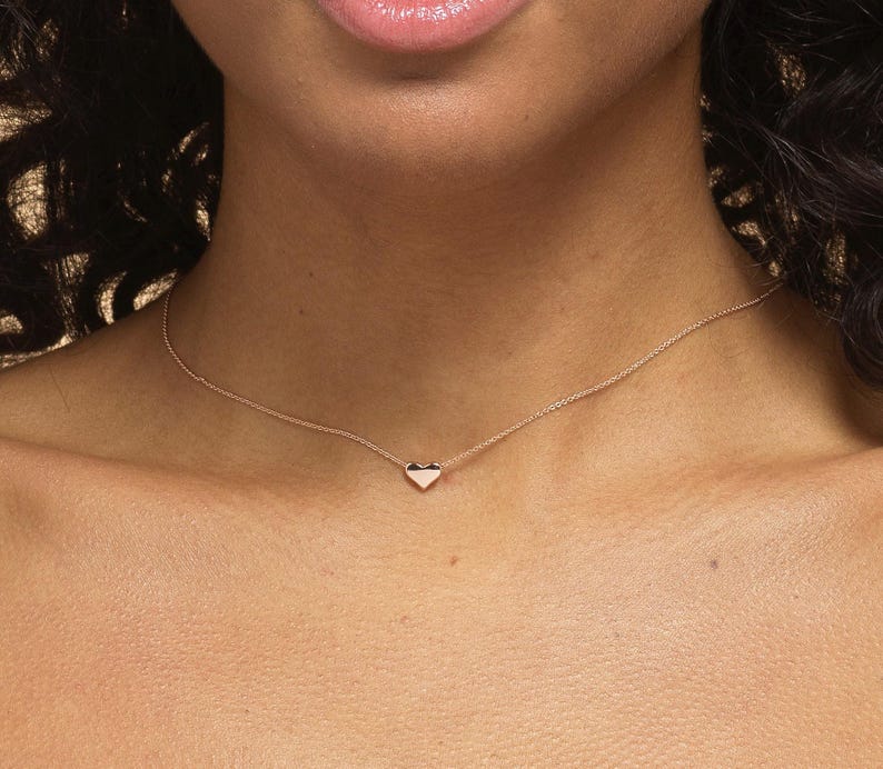 K&ouml;nnte beinhalten: Eine zarte goldene Halskette mit einem kleinen Herzanh&auml;nger. Der herzf&ouml;rmige Anh&auml;nger ist auf einer d&uuml;nnen Kette zentriert, wodurch ein minimalistisches und elegantes Design entsteht. Die Halskette wird um den Hals getragen.