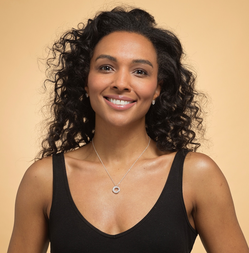 May include: A silver necklace featuring a small, circular pendant with a textured, sparkling surface, suspended from a delicate silver chain. The pendant consists of a single ring design that rests against the chest, providing a subtle metallic accent.