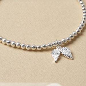May include: A silver bracelet featuring small, round beads and a charm of two angel wings. The wings charm is detailed and shiny. The bracelet is a delicate piece of jewellery, suitable for everyday wear.