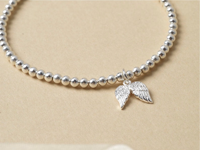 May include: A silver bracelet featuring small, round beads and a charm of two angel wings. The wings charm is detailed and shiny. The bracelet is a delicate piece of jewellery, suitable for everyday wear.
