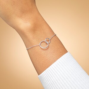 May include: A delicate silver-toned chain bracelet featuring two interlocking circular rings at the centre. The bracelet is constructed with a fine link chain that connects to each side of the two connected rings, creating a simple and continuous design.