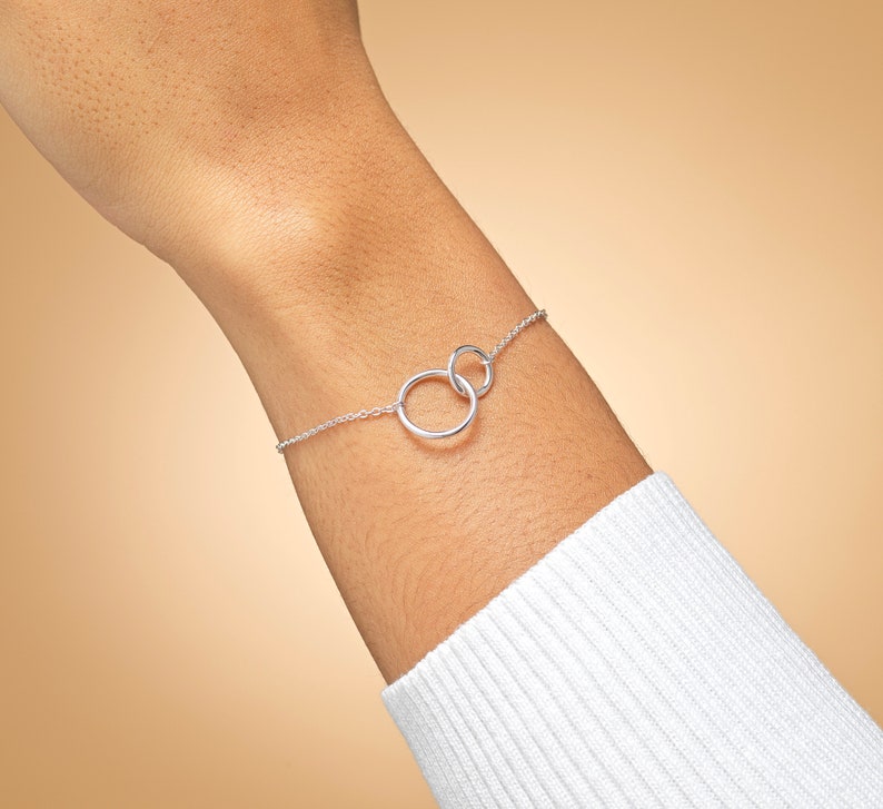 May include: A delicate silver-toned chain bracelet featuring two interlocking circular rings at the centre. The bracelet is constructed with a fine link chain that connects to each side of the two connected rings, creating a simple and continuous design.
