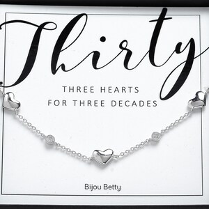 May include: Silver necklace with three heart charms and two clear crystal accents. The necklace is displayed in a box with the text "Thirty" and "Three Hearts for Three Decades" and the brand name "Bijou Betty".