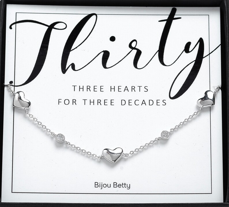 May include: Silver necklace with three heart charms and two clear crystal accents. The necklace is displayed in a box with the text "Thirty" and "Three Hearts for Three Decades" and the brand name "Bijou Betty".