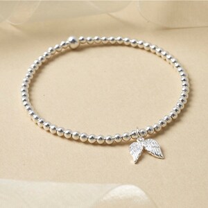 May include: Silver beaded bracelet featuring a charm of two angel wings. The bracelet is composed of small, round, silver beads. The wings charm is detailed, adding a delicate touch to the jewellery.