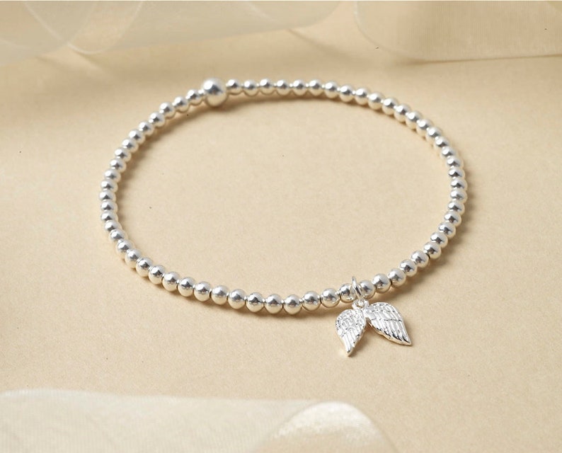 May include: Silver beaded bracelet featuring a charm of two angel wings. The bracelet is composed of small, round, silver beads. The wings charm is detailed, adding a delicate touch to the jewellery.