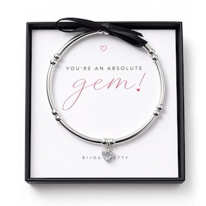 May include: A silver-toned beaded stretch bracelet with a heart-shaped charm, presented in a black gift box. The display card features the text "YOU'RE AN ABSOLUTE gem!" in black and pink script, accented with a small black ribbon bow at the top.