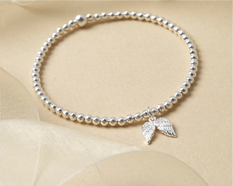 May include: A silver beaded bracelet featuring a charm of two angel wings. The bracelet is composed of small, round, silver beads. The wings charm has etched detailing. The bracelet is presented on a cream-colored, silky surface.