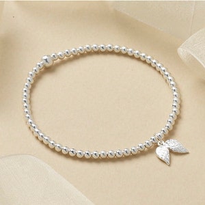 May include: Silver beaded bracelet featuring a charm of two small angel wings. The bracelet is made of small, round, silver beads. The wings are detailed, adding a delicate touch to the jewellery.