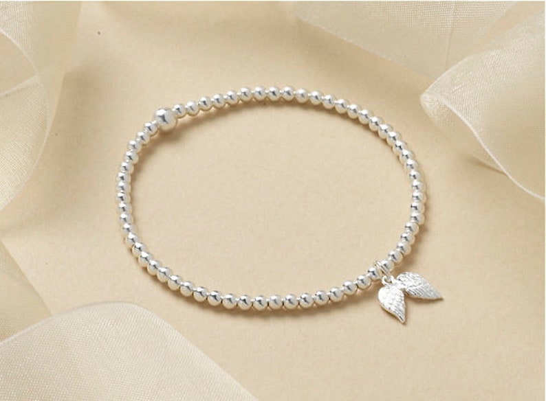 May include: Silver beaded bracelet featuring a charm of two small angel wings. The bracelet is made of small, round, silver beads. The wings are detailed, adding a delicate touch to the jewellery.