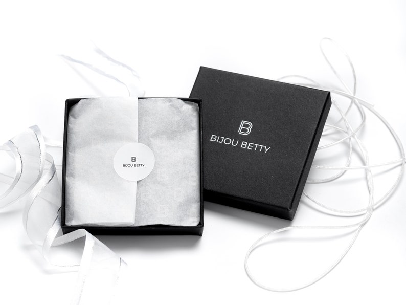 May include: A black gift box with the text "BIJOU BETTY" in white. The box is open, revealing white tissue paper and a round sticker with the "BIJOU BETTY" logo. White ribbon is draped around the box.